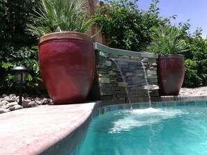 Une piscine agrémentée d’une cascade, entourée de grands bacs à fleurs rouges et d’un mur de pierre.
