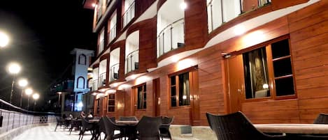 A building with multiple floors, outdoor seating area with tables and chairs, and illuminated windows.
