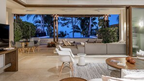Una sala de estar moderna con un televisor grande, zona de estar y vista a palmeras al atardecer.