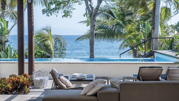 Un área de descanso junto a la piscina con vista al océano, sillones de playa y una mesita con cojín.