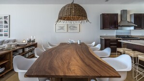 Una cocina moderna con una mesa de madera larga, sillas blancas, armarios oscuros y una lámpara colgante de fibras naturales.