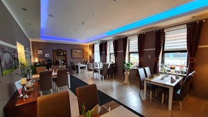 Comedor de hotel con varias mesas y sillas, un ventanal con cortinas e iluminación empotrada en el techo.