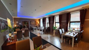 Comedor de hotel con varias mesas y sillas, un ventanal con cortinas e iluminación empotrada en el techo.
