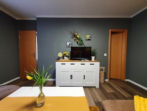 Un salon avec un téléviseur à écran plat posé sur un meuble blanc, un vase rempli de fleurs et une table basse en bois.