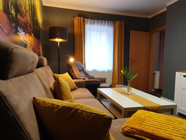 A modern living room with a sofa, armchair, and a coffee table. There is a lamp, a window with curtains, and a radiator.