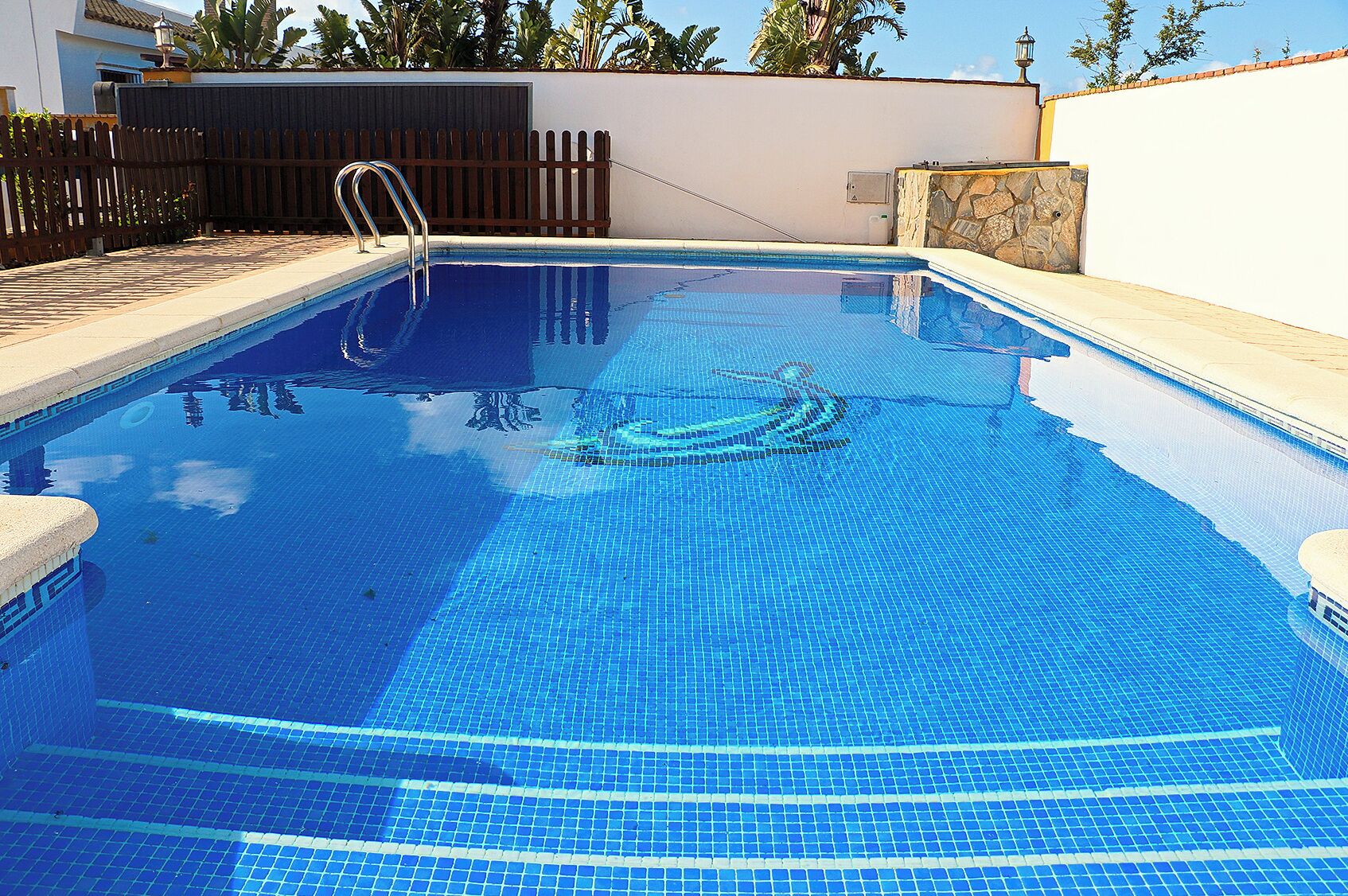 Una piscina rectangular amb aigua blava i transparent, envoltada d'una terrassa amb rajoles i una tanca de fusta.