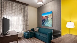 A modern hotel room with a sectional sofa, a flat-screen TV, a small table, a lamp, and a painting of a canyon.