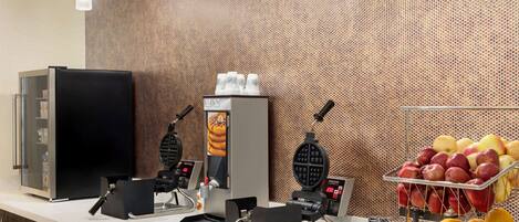 A breakfast bar with waffle makers, a refrigerator, and a fruit display.