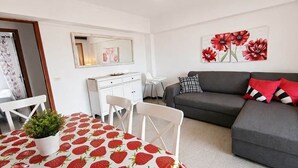 Un salon avec un canapé gris, une table à manger recouverte d’une nappe à motifs de fraises, une commode blanche et un miroir au mur.