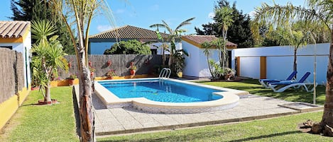 Una piscina rectangular con una solana con baldosas, rodeada por un césped cuidado y diversas plantas en macetas.