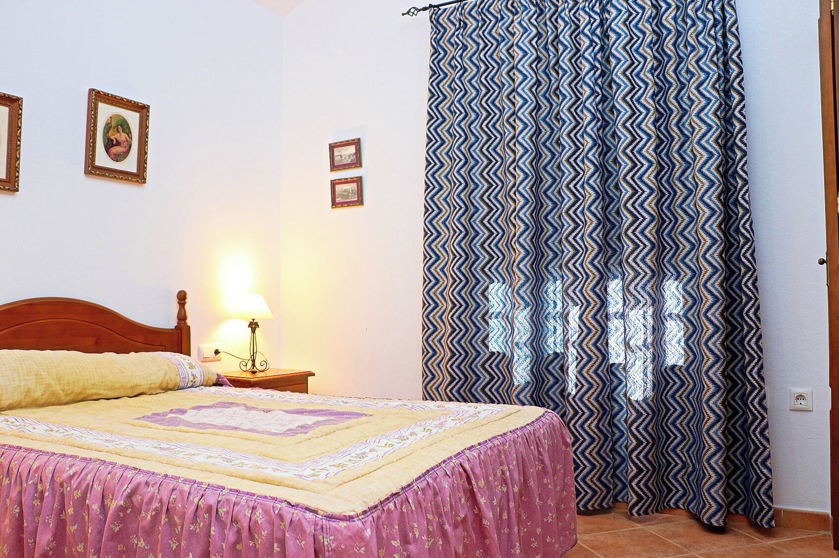 Une chambre à coucher avec un lit, une table de chevet avec une lampe, un rideau à motifs et des tableaux encadrés accrochés au mur.