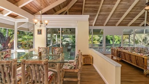 A spacious room with a high ceiling and wooden beams, featuring a glass dining table with chairs, a sofa, and a view of lush greenery outside. - Peaceful Location with Lush Tropical Views (Princeville)