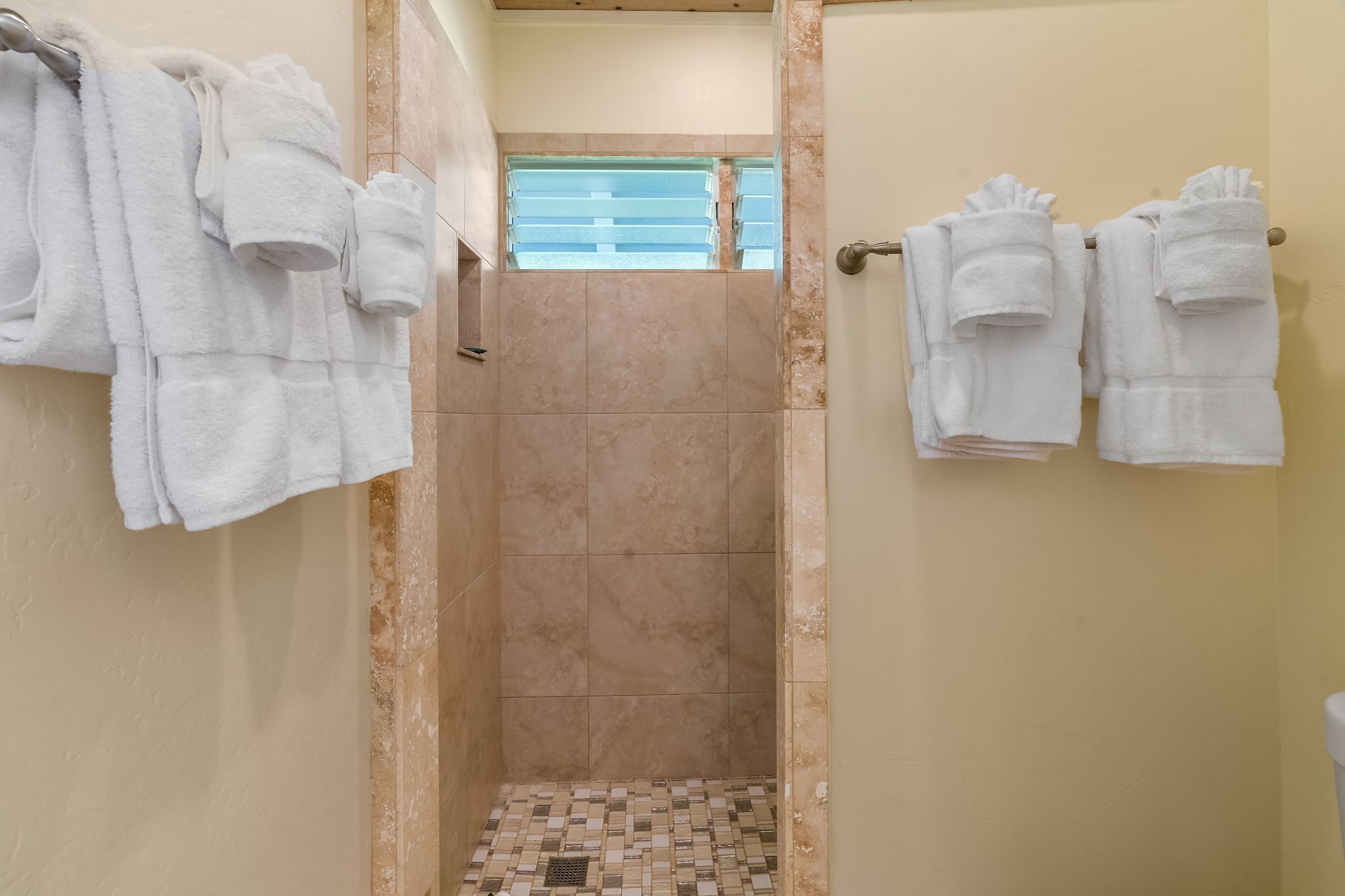 A bathroom with a tiled shower area, beige walls, and white towels hanging on the wall.