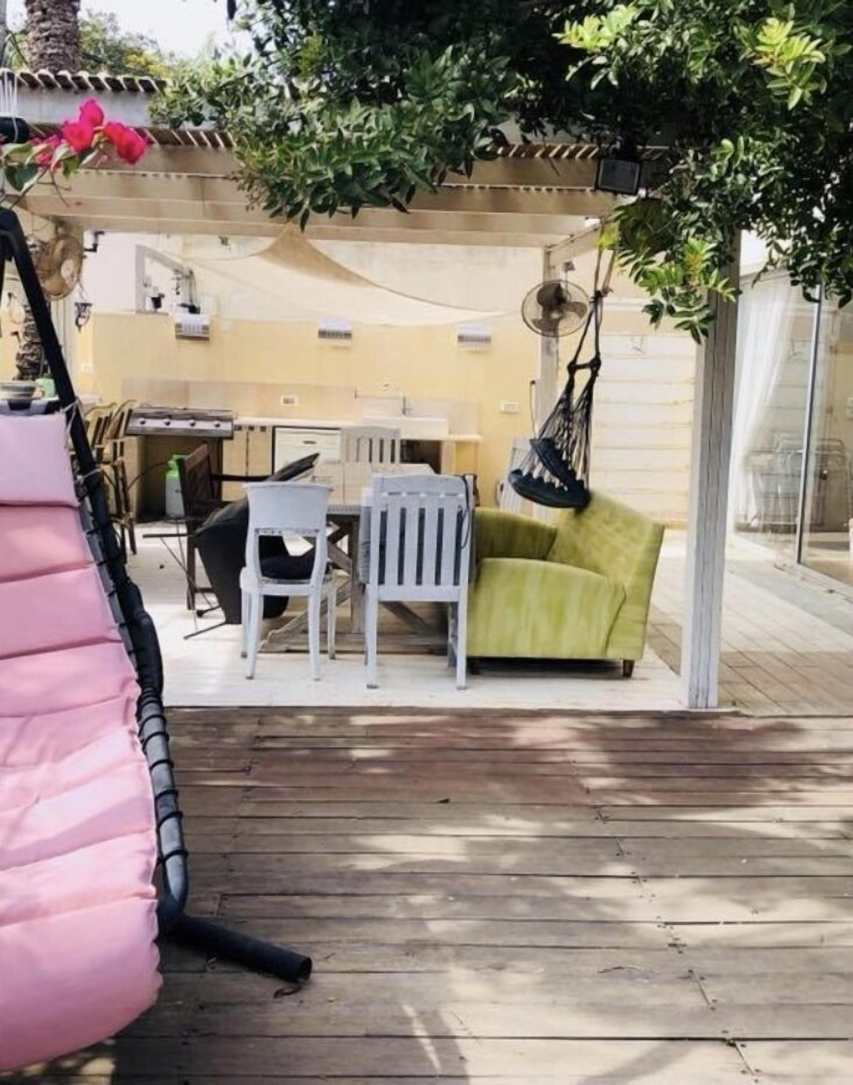 Un patio exterior con piso de madera, una mesa blanca con sillas, un sofá verde y una silla colgante.