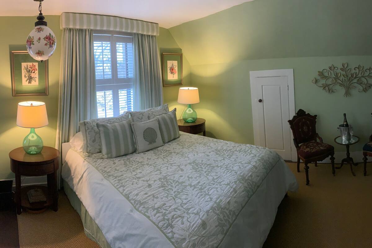 A bedroom with a large bed, two bedside tables with lamps, a window with curtains, a door, a chair, a small table with a bottle, and a vase with flowers.