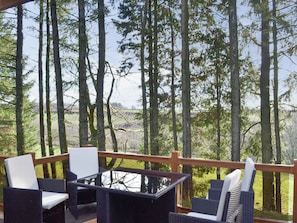 A covered outdoor seating area with wicker furniture and a glass table, overlooking a forested landscape.