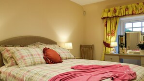 Une chambre à coucher avec un lit, des oreillers, une table de chevet avec une lampe, un miroir et une fenêtre avec des rideaux.