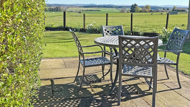 Une terrasse avec une table et des chaises, une haie et une vue sur un champ verdoyant et des collines lointaines.