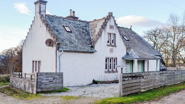 Een wit, tweelaags gebouw met een flauwe dakschild en een schoorsteen, en een klein raam met de tekst 'DAIRY COTTAGE'.