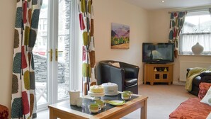 A living room with a sofa, a television, a table with a cake and cups, and a window with curtains. - 3 bedroom accommodation in Ambleside (Ambleside)