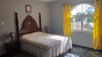 A bedroom with a wooden bed, two bedside tables with lamps, a ceiling fan, a window with yellow curtains, and a patterned floor.