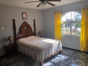 A bedroom with a wooden bed, two bedside tables with lamps, a ceiling fan, a window with yellow curtains, and a patterned floor.