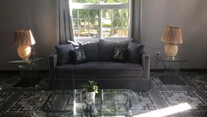 A modern living room with a dark grey sofa, two lamps, a glass coffee table, and a large window with curtains. - Retreat Comfortable Apartments (Falmouth)
