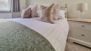 Een net opgemaakt bed met een groene deken, een nachtkastje met een lamp en een raam met gordijnen.