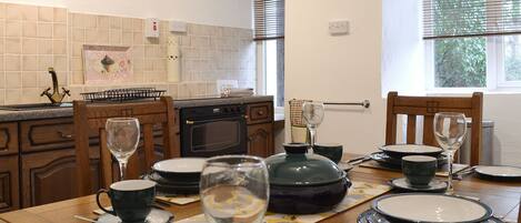 A dining area with a wooden table set for a meal, a kitchen in the background, and a clock on the wall.