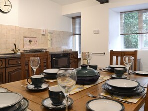 Un comedor con una mesa de madera dispuesta para una comida, una cocina al fondo y un reloj en la pared.
