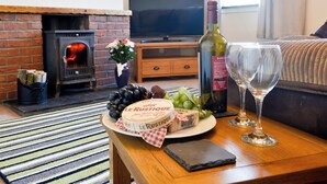 Un salon chaleureux avec une cheminée, un téléviseur, une table sur laquelle on trouve un plateau de fromages, des raisins, une bouteille de vin et deux verres à vin.