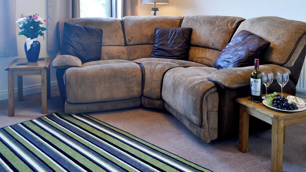A corner living room with a sectional sofa, a small side table with a vase of flowers, a lamp, and a table with a bottle of wine and grapes. - 2 bedroom accommodation in Mabie, near Dumfries (Mabie, near Dumfries)