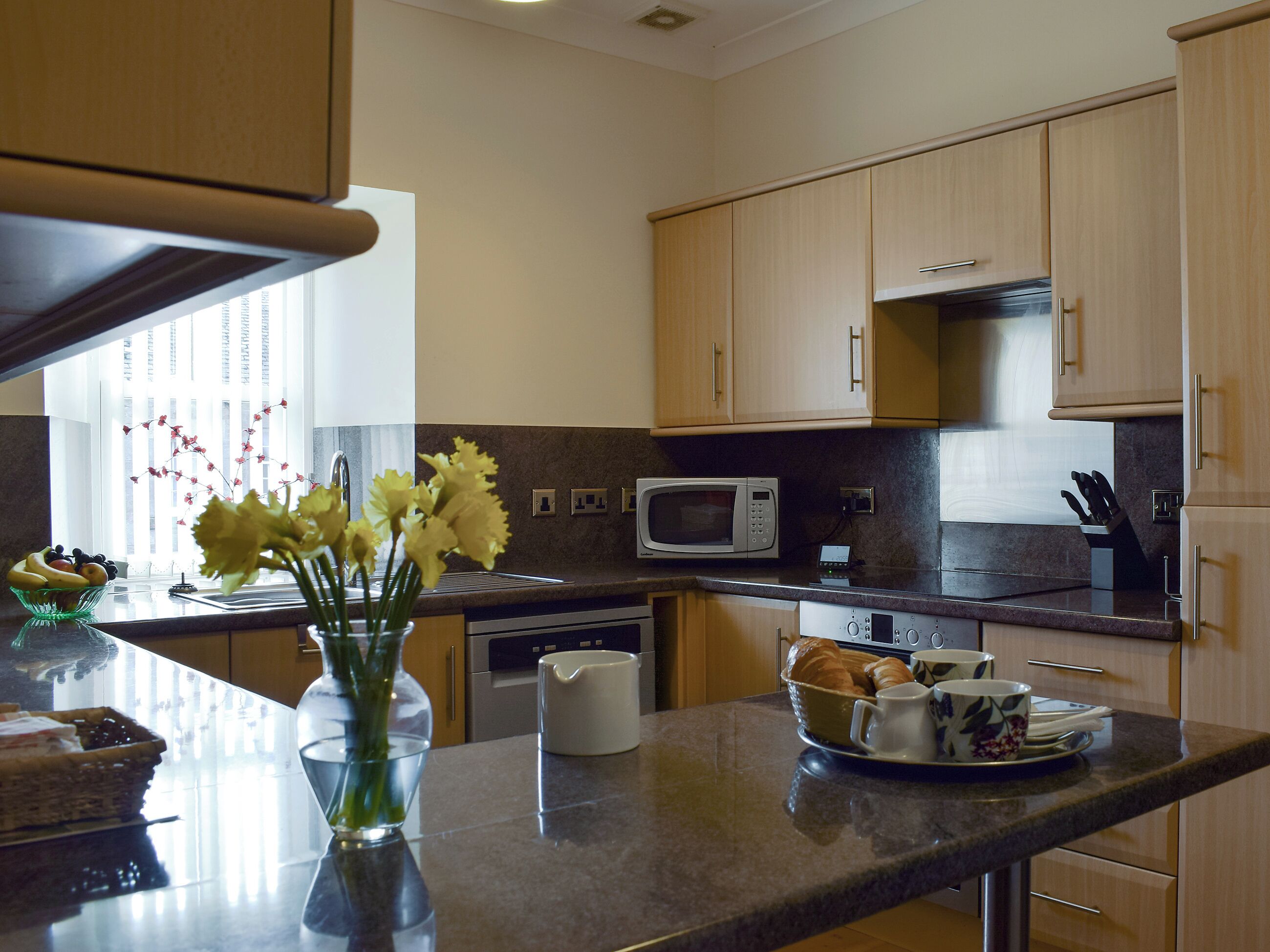 A modern kitchen with wooden cabinets, a microwave, an oven, and a countertop with a vase of flowers and a fruit bowl.