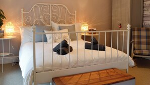 Una cama bien hecha con cabecera blanca, una mesita de noche con lámpara y un banco de madera en una habitación con una paleta de colores neutros.