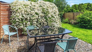 A patio with a table and chairs, surrounded by a garden with a large flowering bush. - 2 bedroom accommodation in Derril, near Bude (Derril, near Bude)