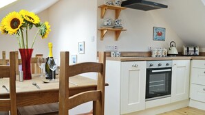 Une cuisine avec des meubles de rangement en bois, un four encastré, une table à manger en bois avec des chaises, un vase rouge contenant des tournesols et une bouteille de vin.