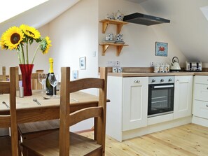 A kitchen with wooden cabinets, a built-in oven, a wooden dining table with chairs, a red vase with sunflowers, and a wine bottle.