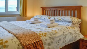Un lit bien fait, avec du linge de lit blanc et un plaid à motifs, une tête de lit en bois et une vue sur la mer par la fenêtre.