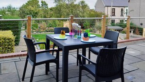 Une terrasse extérieure avec une table dressée pour deux personnes, entourée de chaises en osier, et une vue sur un jardin et un bâtiment en arrière-plan.