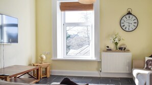 A living room with a sofa, a wooden table, a globe, a window with a view of trees, a radiator, a clock, and a television on the wall. - 3 bedroom accommodation in Keswick (Keswick)