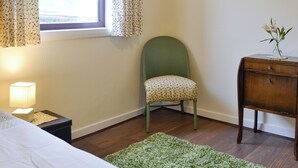 A bedroom with a bed, a nightstand, a green chair, a wooden dresser, a window with curtains, and a framed picture on the wall.