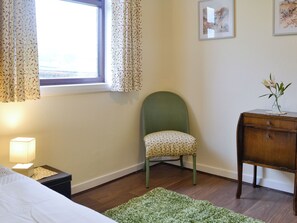 A bedroom with a bed, a nightstand, a green chair, a wooden dresser, a window with curtains, and a framed picture on the wall.