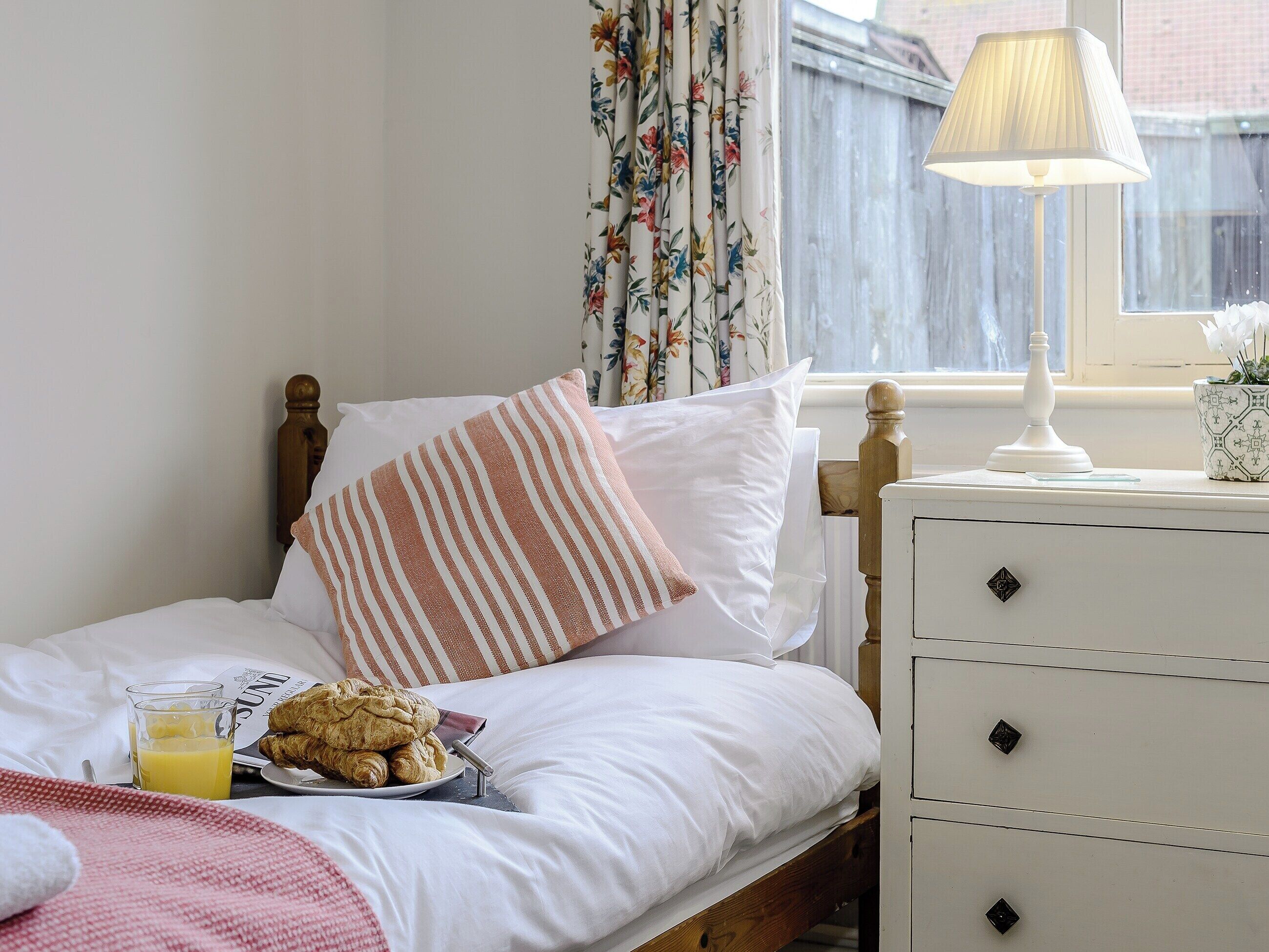 A neatly made bed with a striped pillow, a bedside table with a lamp, and a plate of croissants and orange juice on the nightstand.