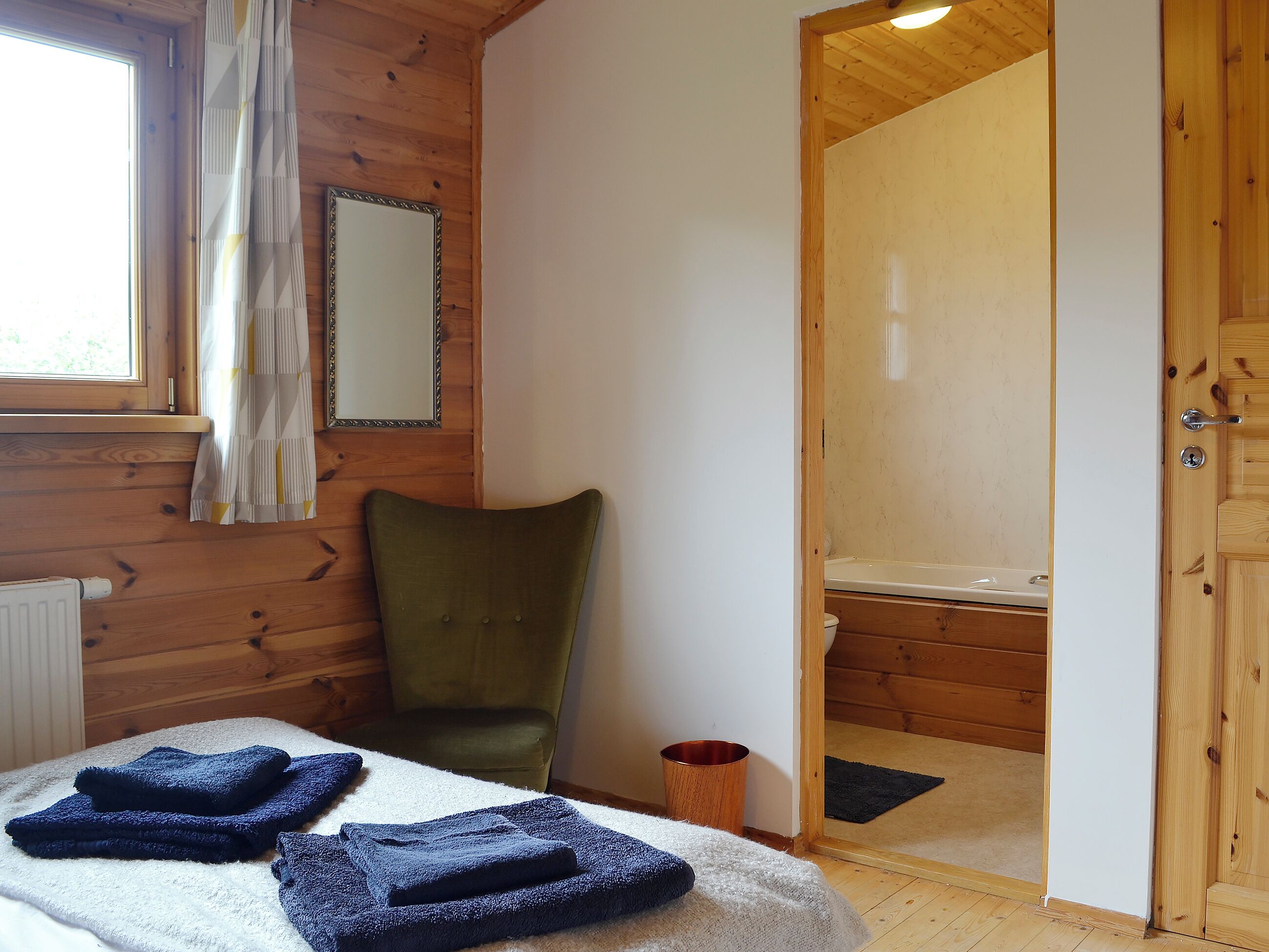 A wooden-paneled room with a bed, a green chair, and a bathroom visible through an open door.