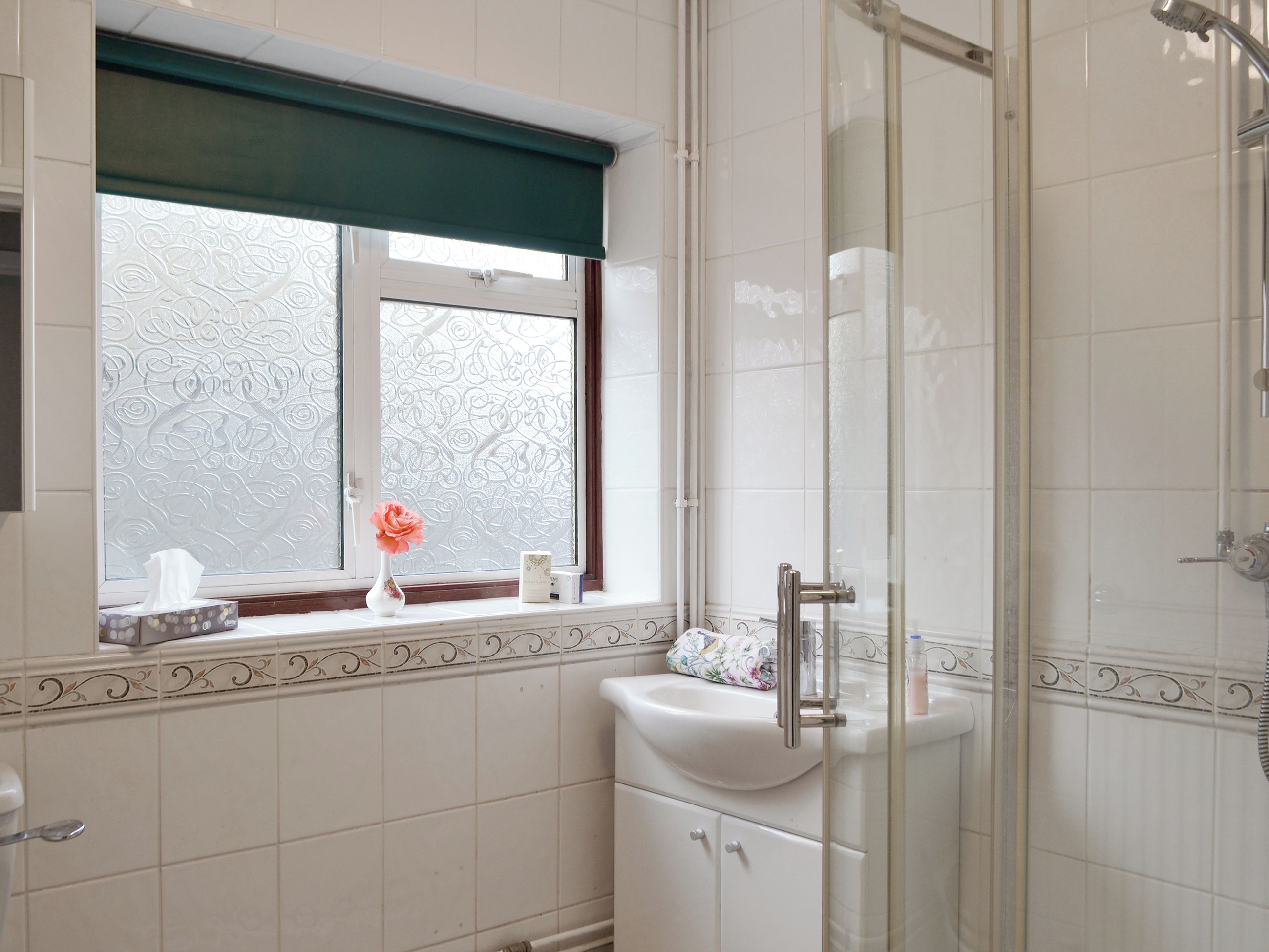 Ein Badezimmer mit Duschkabine aus Glas, einem Fenster mit Zierrollo und einem Waschbecken mit Blumen in einer Vase.