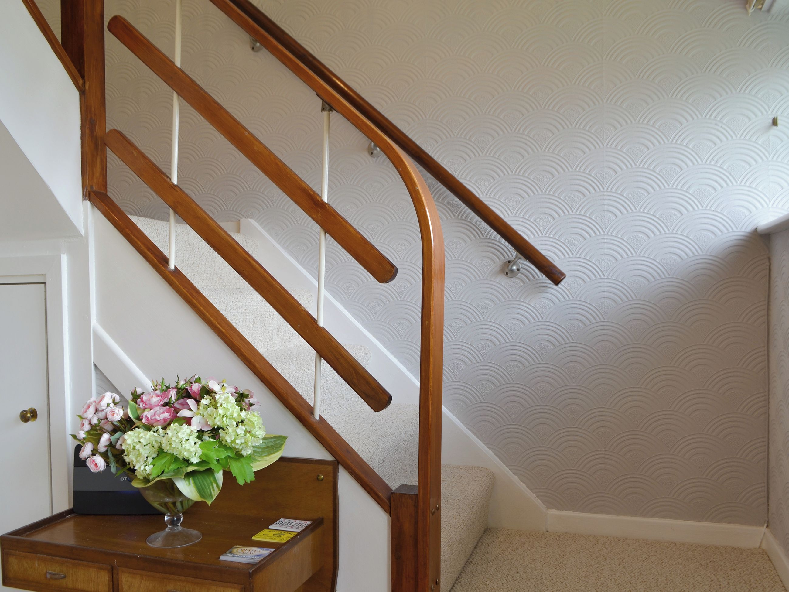 A wooden staircase with a handrail, a wall with a textured pattern, and a desk with a vase of flowers.