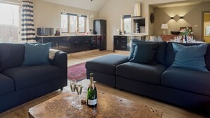 Ein modernes Wohnzimmer mit dunklen Sofas, einem Couchtisch mit einer Flasche und Gläsern sowie einer Küche im Hintergrund.