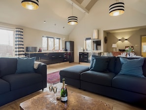 A modern living room with dark sofas, a wooden coffee table with a bottle and glasses, and a kitchen in the background. - 3 bedroom accommodation in Stretton-on-Fosse, near Moreton-in-Marsh (Stretton-on-Fosse, near Moreton-in-Marsh)