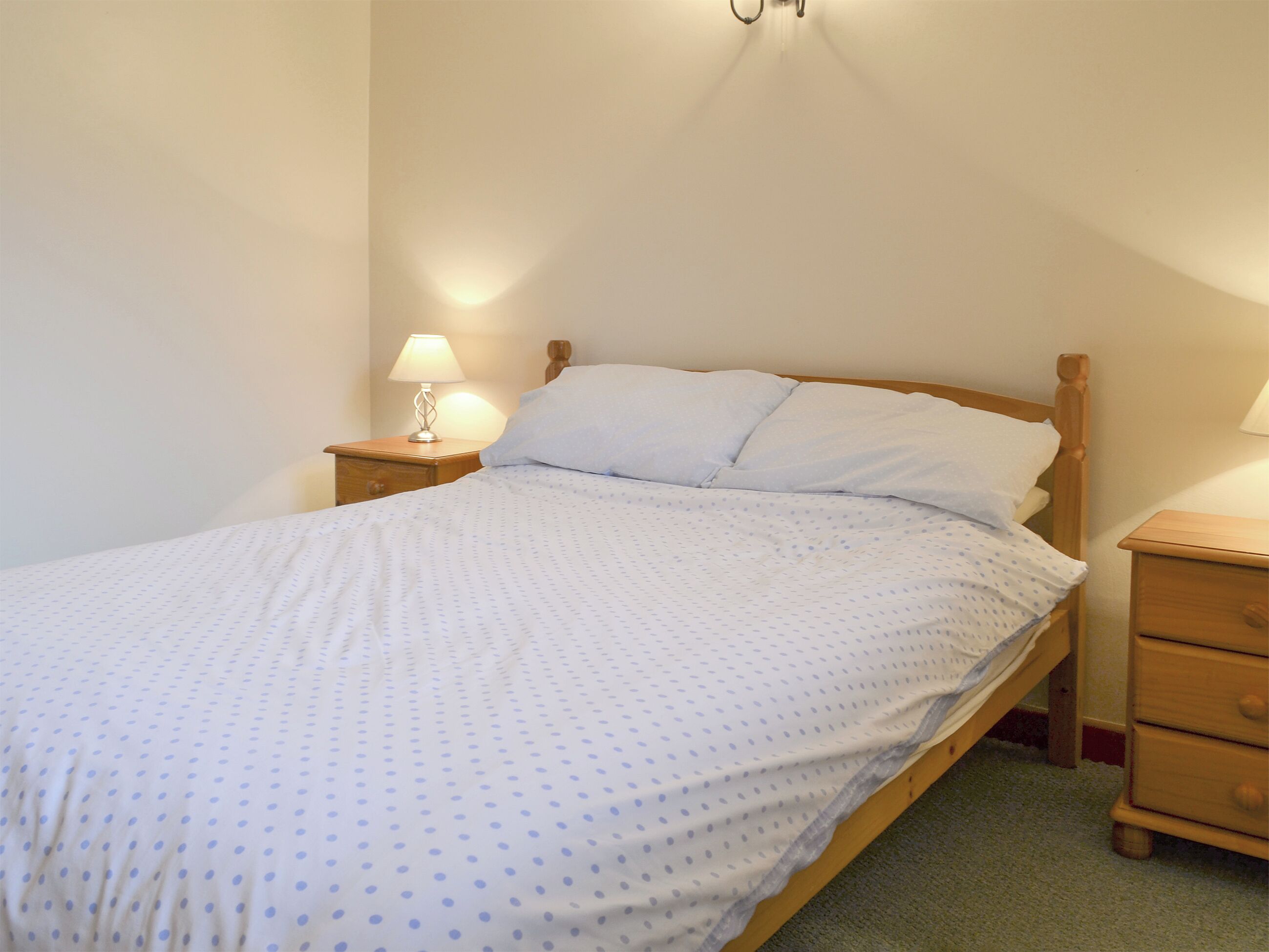A single bed with a wooden headboard and footboard, a white bedspread with blue polka dots, two bedside tables with lamps, and a wooden nightstand with a drawer.