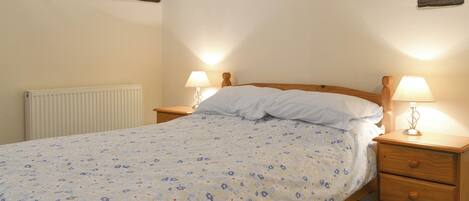 Quarto com cama, duas mesas de cabeceira com luminárias, cabeceira de madeira, radiador e teto inclinado com vigas aparentes.